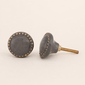 May include: Two gray ceramic cabinet knobs with gold-colored accents. The knobs have a round shape with a decorative border of small gold dots. Each knob has a threaded brass stem for installation.