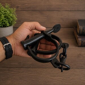 May include: A black metal shackle featuring a circular cuff and a locking pin mechanism. The device is constructed from heavy-duty iron with a smooth, dark finish, consisting of a hinged ring connected to a short chain and a T-shaped locking bolt.