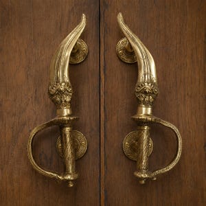 May include: A pair of polished brass door handles shaped like curved swords or daggers. Each handle features an ornate hilt with a textured grip, a curved guard, and a blade-like upper section with intricate engraved details and circular mounts.