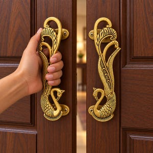 May include: A pair of gold-coloured metal door handles featuring a detailed peacock design with intricate feather patterns. Each handle is mounted vertically onto a dark brown wooden door, with the peacock head positioned at the bottom.