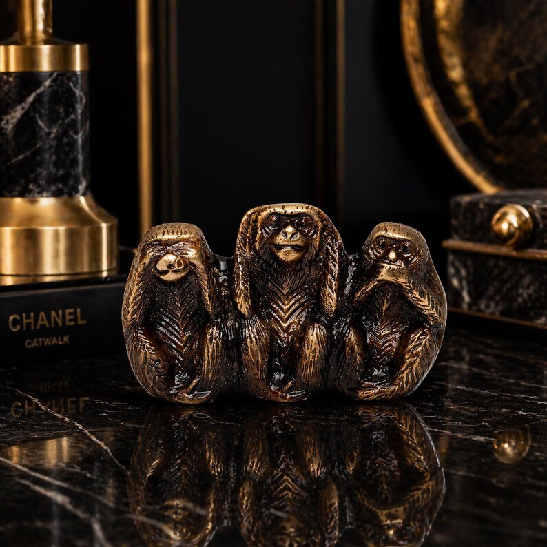 May include: A bronze-colored sculpture of the three wise monkeys, representing "see no evil, hear no evil, speak no evil." The monkeys are in a row, with one covering its eyes, one its ears, and one its mouth. The sculpture is on a black surface.