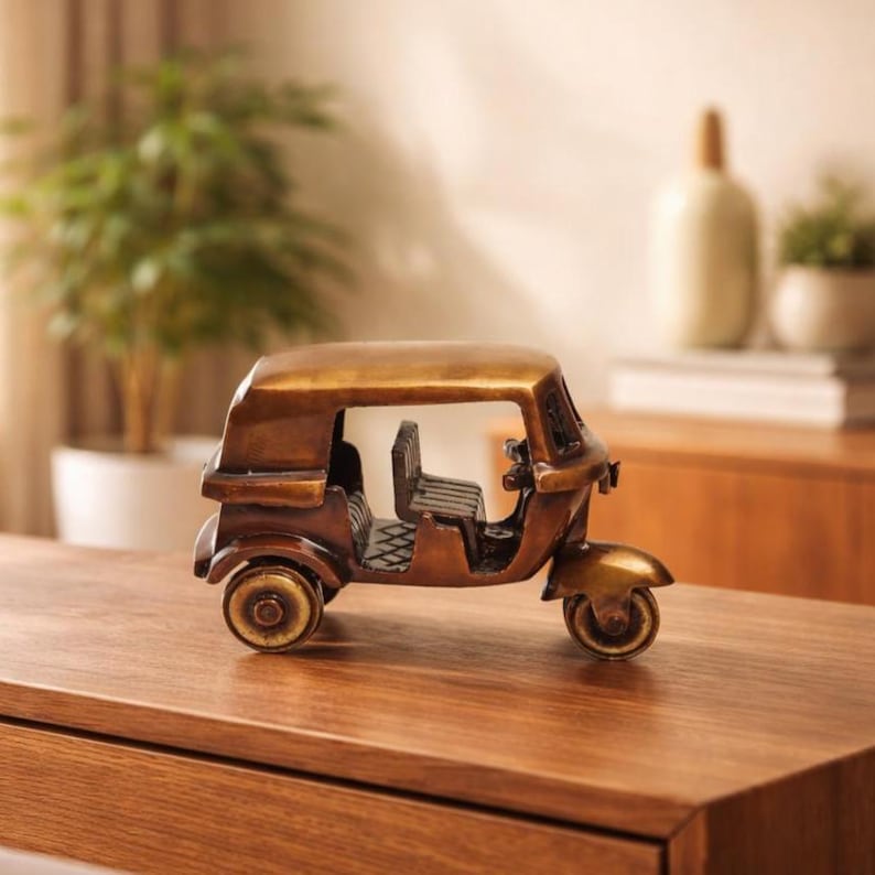 May include: A bronze-coloured decorative auto-rickshaw model. The miniature vehicle features a detailed design with a roof, seats, and wheels. The model is displayed on a wooden surface, showcasing its intricate craftsmanship and vintage aesthetic.