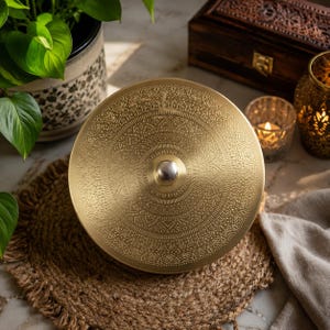May include: A circular brass container lid featuring an intricate, embossed mandala-style pattern across its surface. The lid has a polished, metallic gold finish and a small, smooth, silver-toned knob centered at the top for easy lifting.