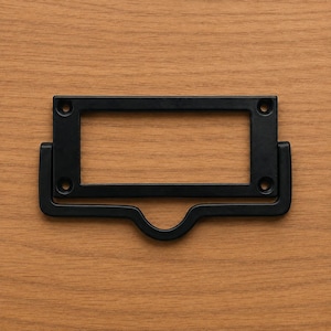 Op de afbeelding: Een matzwarte metalen labelhouder met een rechthoekig frame voor het plaatsen van labels en een gebogen handgreep eronder. Het beslag is voorzien van vier ronde schroefgaten op de hoeken van het frame voor een stevige bevestiging.