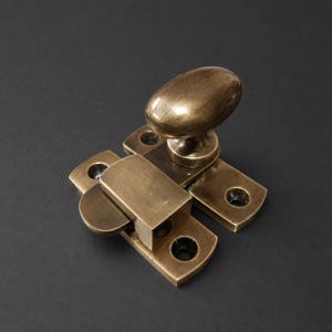 Peut inclure: Un verrou de fenêtre en laiton doté d'une poignée ovale et d'une plaque de base rectangulaire avec quatre trous de vis pré-percés. La quincaillerie métallique présente une finition brossée et vieillie avec des tons métalliques visibles.