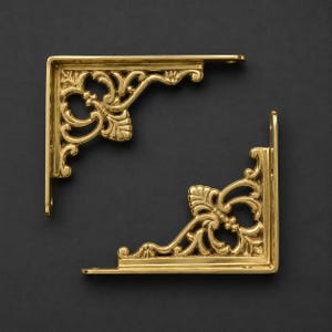 May include: Two L-shaped shelf brackets made of polished gold-toned metal. Each bracket features an intricate, ornate scrollwork design with leaf-like motifs filling the corner space. Both brackets have flat mounting arms with visible screw holes.