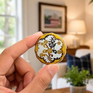 May include: A round ceramic knob featuring a white center with a dark brown and mustard yellow floral pattern. The edges of the knob are scalloped and finished with a mustard yellow border, creating a decorative accent for furniture or cabinetry.