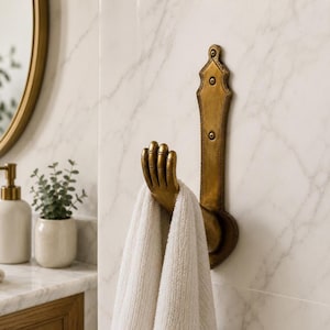 May include: A wall-mounted brass-toned metal hook featuring a sculpted hand design. The vertical backplate is secured with two visible screws, and a white textured towel is draped over the fingers of the hand-shaped hook for display.