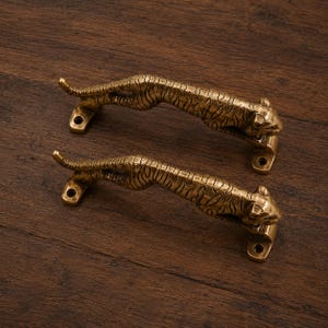 May include: Two matching brass-toned metal door handles shaped like leaping tigers. Each handle features a textured, striped body with a head at one end and a tail at the other, including two circular mounting plates with pre-drilled screw holes.
