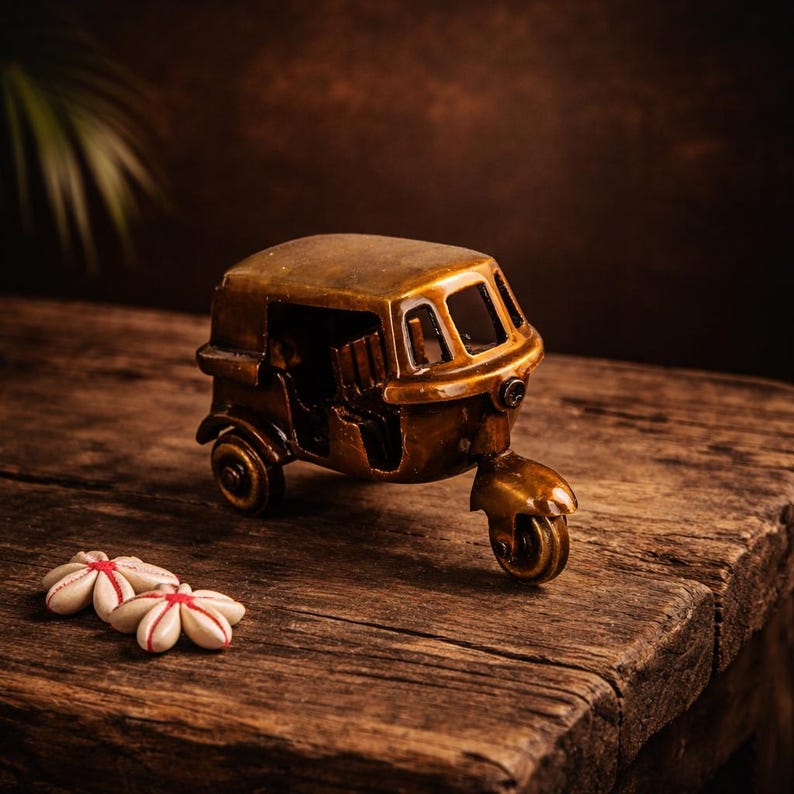 May include: A bronze-toned miniature auto rickshaw model, a three-wheeled vehicle, is displayed on a rustic wooden surface. The model features detailed wheels and a driver's area. Two small, white flower decorations with red detailing are also visible.