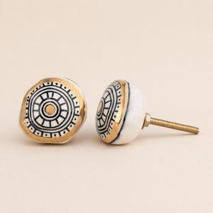 May include: Two decorative cabinet knobs. The knobs are white with a gold rim and a black and gold circular design. One knob is positioned on its side, revealing the threaded screw.