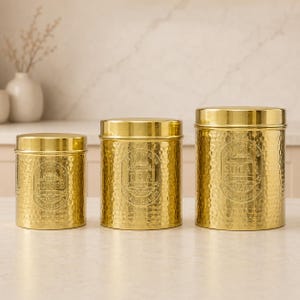 Op de afbeelding: Een set van drie cilindervormige goudkleurige metalen bussen met een gehamerde textuur en bijpassende deksels. Elke bus heeft een reliëflabel met decoratieve randen en de tekst Coffee, Tea en Sugar.