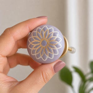 Puede incluir: Un pomo de armario de cerámica lavanda con un diseño floral dorado y blanco. El pomo tiene forma redonda y una base de latón. El patrón floral está centrado en la cara del pomo. El pomo se sostiene en una mano.