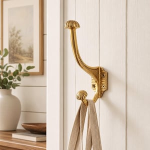 May include: A polished gold-tone metal wall hook featuring a double-prong design with a curved upper arm and a smaller lower hook, both topped with rounded, ribbed finials. The base plate is rectangular with four visible screw mounting holes.