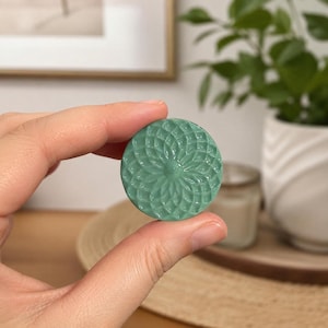 May include: A small, circular, sage green object featuring a raised, symmetrical geometric pattern of overlapping petals or leaves radiating from a central point. The surface has a smooth, matte finish with intricate, repeating floral-like textures.