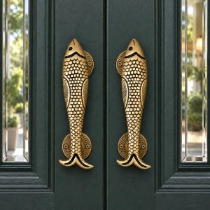 Puede incluir: Un par de manijas de puerta verticales de latón con forma de pez, que presentan texturas de escamas y aletas detalladas. Cada manija está montada en una puerta de color verde oscuro con placas circulares en los extremos y tornillos visibles.