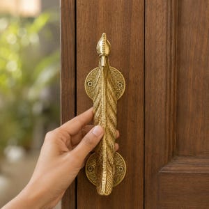 May include: A gold-toned metal door handle shaped like a stylized feather with intricate textured details. The handle is mounted vertically onto a dark brown wooden door with two circular base plates, each secured by visible screws at the top and bottom.