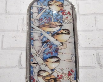 May include: A silver-toned pendant necklace with an oval-shaped pendant. The pendant features a detailed illustration of several small birds perched on branches with red berries against a blue and white background. The necklace has a delicate chain.