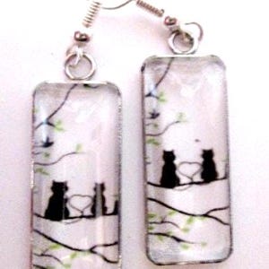 May include: A pair of rectangular drop earrings featuring a white background with a black silhouette illustration of two cats sitting on a tree branch. The design includes thin black branches with small green leaves and a heart shape between the cats.