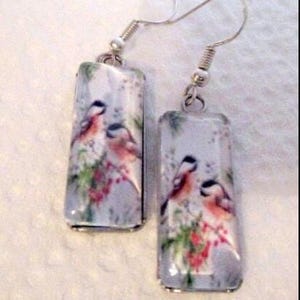 May include: A pair of rectangular dangle earrings. Each earring features a detailed illustration of two birds perched on a branch with red berries and green leaves. The earrings have silver-colored hooks.