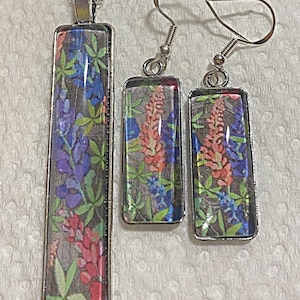 May include: A silver-toned jewelry set consisting of a long rectangular pendant necklace and matching rectangular drop earrings. Each piece features a vibrant floral print with blue, pink, and green lupine flowers set under a clear glass cabochon.