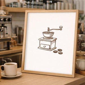 May include: A framed illustration of a coffee grinder with coffee beans. The drawing is in brown ink on a white background. The frame is light brown. A coffee cup and espresso machine are in the background.