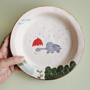 May include: A ceramic plate with a whimsical illustration of a gray elephant holding a red umbrella in the rain. The plate has a cream-colored base with a brown rim and green and blue painted accents.