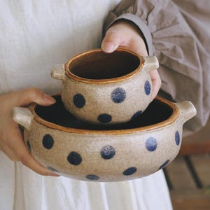 May include: Two round ceramic bowls with handles, featuring a speckled beige finish and dark blue polka dot patterns. The smaller bowl is nested inside the larger bowl, both showing a smooth interior rim and a textured, earthy exterior surface.