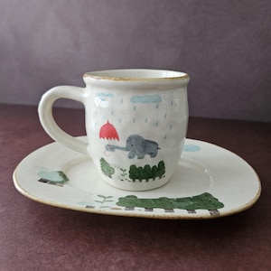 May include: A ceramic mug and matching oval saucer featuring a hand-painted scene of a gray elephant holding a red umbrella under blue rain clouds. The off-white pottery has a speckled texture and a brown rim detail on both pieces.