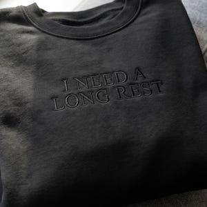 May include: A black crewneck sweatshirt featuring the embroidered text I NEED A LONG REST in a matching black thread. The garment has a ribbed collar, long sleeves, and a clean, solid-colored design with centered lettering on the chest area.