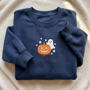 May include: A navy blue crewneck sweatshirt featuring a central embroidered design of an orange jack-o'-lantern pumpkin, a small white ghost, and several cream-colored stars arranged around the figures on the front of the garment.