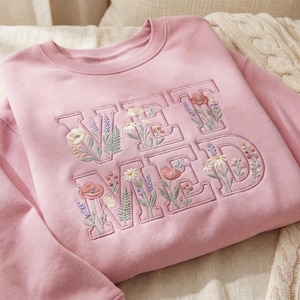 May include: A light pink crewneck sweatshirt featuring the embroidered text VET MED in a bold block font. The letters are filled with detailed floral embroidery, including various wildflowers, poppies, and lavender sprigs in soft pastel colors.