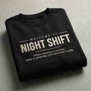 May include: A black crewneck sweatshirt featuring embroidered cream-colored text that reads: WELCOME TO NIGHT SHIFT, followed by the smaller text: Where everyday is monday, today is yesterday, and tomorrow is today, framed by two horizontal lines.