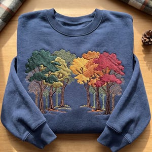 Puede incluir: Una sudadera azul de cuello redondo con un dise&ntilde;o bordado de un bosque. El bordado muestra &aacute;rboles con hojas en tonos verdes, amarillos, naranjas y rojos, representando una transici&oacute;n del follaje de verano a los colores del oto&ntilde;o.