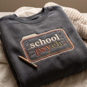 May include: A dark gray sweatshirt with the embroidered text "school psych" in cream, green, and orange. The design is framed by a stitched border and a paperclip. A wooden pencil rests on the fabric.