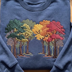 Puede incluir: Una sudadera azul de cuello redondo con un dise&ntilde;o bordado detallado de una escena forestal. El bordado muestra varios &aacute;rboles con follaje en tonos verde oscuro, verde claro, amarillo, naranja y rojo sobre la tela azul s&oacute;lida.