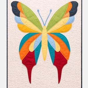 Op de afbeelding: Een kleurrijke vlinderquilt met een crèmekleurige achtergrond. De vlinder heeft vleugels in groen, geel, oranje, blauw en rood. De quilt is omlijst met een donkere rand en heeft een gewatteerd patroon.