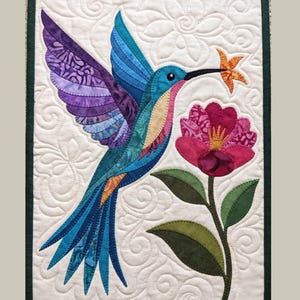 May include: A rectangular fabric wall hanging featuring a quilted white background with swirling patterns. A colorful hummingbird in shades of purple, blue, and orange hovers near a large pink flower with green leaves, all crafted from fabric.