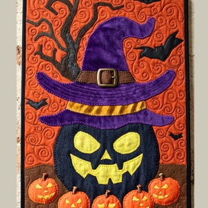 May include: A rectangular fabric wall hanging featuring a large black jack-o'-lantern wearing a purple witch hat with a brown buckle. The background is orange with black swirling patterns, silhouettes of bats, and a tree. Five small orange pumpkins sit below.