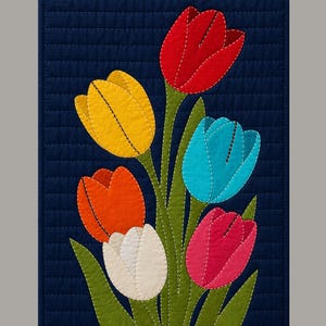 Può includere: Un arazzo trapuntato con un bouquet di tulipani colorati. I tulipani sono nei toni del rosso, giallo, arancione, blu, rosa e bianco, con steli e foglie verdi. Lo sfondo è un tessuto blu scuro con cuciture orizzontali.