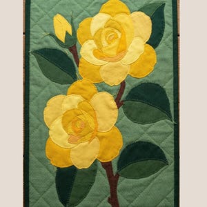 May include: A quilted wall hanging with two yellow roses, green leaves, and a bud on a green background. The piece is framed with a dark green border and has a diamond pattern. The dimensions are approximately 30 cm x 20 cm.