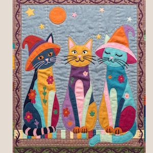 Op de afbeelding: Een kleurrijk, gewatteerd wandkleed met drie grillige katten met heksenhoeden. De katten zijn in tinten blauw, oranje, geel en paars, versierd met bloemmotieven. Een maan en sterren zijn op de achtergrond te zien.