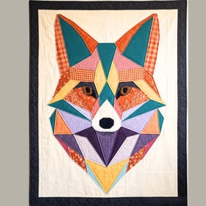 May include: A geometric fox quilt featuring a patchwork design with various fabrics in teal, orange, pink, purple, and yellow. The fox has brown eyes and a black nose, set against a cream background with a black border.