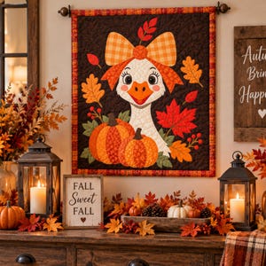 Peut inclure: Une tenture murale matelassée représentant une oie blanche avec un grand nœud à carreaux orange, entourée de citrouilles orange, de feuilles d'automne rouges et orange, et de baies sur un fond marron foncé, bordée d'un tissu à carreaux.