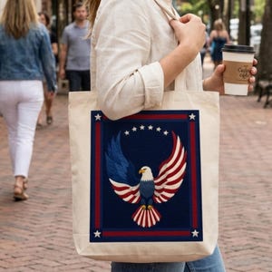 Puede incluir: Un bolso tote de color crema con un dise&ntilde;o patri&oacute;tico. El dise&ntilde;o incluye un fondo azul con un borde rojo y blanco, un &aacute;guila con alas rojas, blancas y azules y seis estrellas blancas. El bolso lo lleva una persona.
