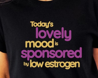 Puede incluir: Camiseta negra con el texto "Today's lovely mood is sponsored by low estrogen" en amarillo y morado.