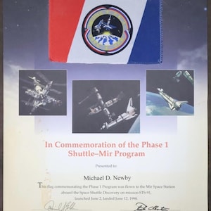 以下が含まれることがあります： 赤、白、青のストライプと円形エンブレムが付いた旗が特徴の、シャトルミールプログラムを記念したポスター。ポスターには、スペースシャトルとミール宇宙ステーションの画像とテキストが含まれています。