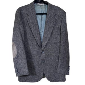 May include: A gray tweed blazer with a notched lapel, two-button closure, and a light blue lining. The blazer features a gray suede elbow patch on the left sleeve. The jacket is hanging on a black hanger.
