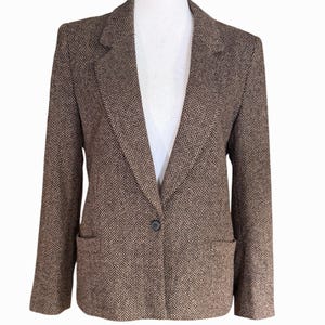Puede incluir: Un blazer marrón y negro con estampado de espiga, que presenta un cierre de un solo botón, solapas de muesca y dos bolsillos delanteros. La prenda tiene mangas largas y un corte estructurado con un patrón tejido clásico.