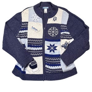 May include: A navy blue zip-up cardigan featuring a patchwork design with cream and light blue panels. The knit sweater displays winter-themed embroidery including snowflakes, a sled, an ice skate, and a mitten, with ribbed trim.